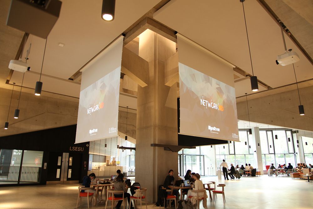 Large open space at LSE with project screens and people seated at tables socialising.